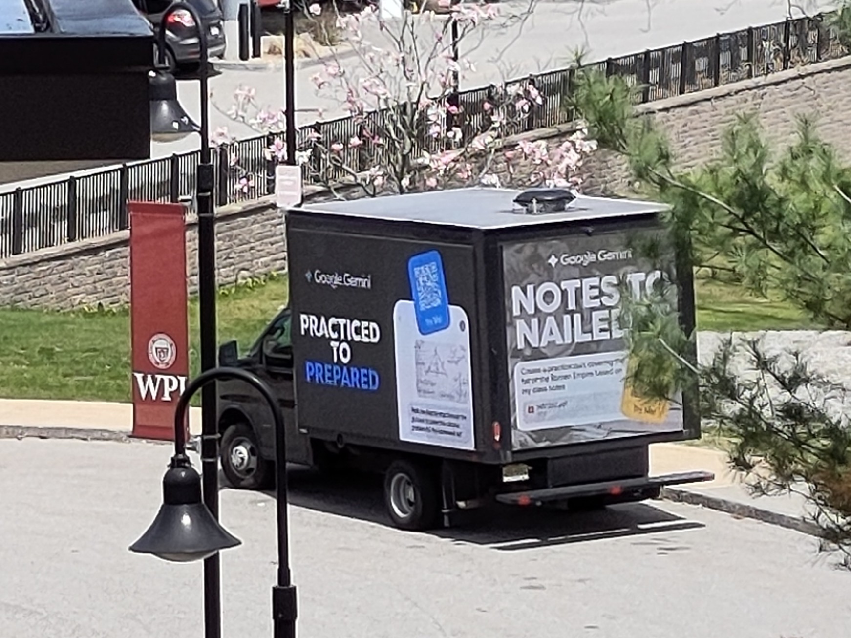 Black mobile billboard truck parked in cul-de-sac behind the WPI Sports and Recreation Center, close to the gravel where the portable classroom used to be. Two advertisements are visible: on the left side, a Google Gemini logo is visible, with a headline below headline reading "PRACTICED TO PREPARED"; and at the back of the truck, a similar ad says "NOTES TO NAILED IT"