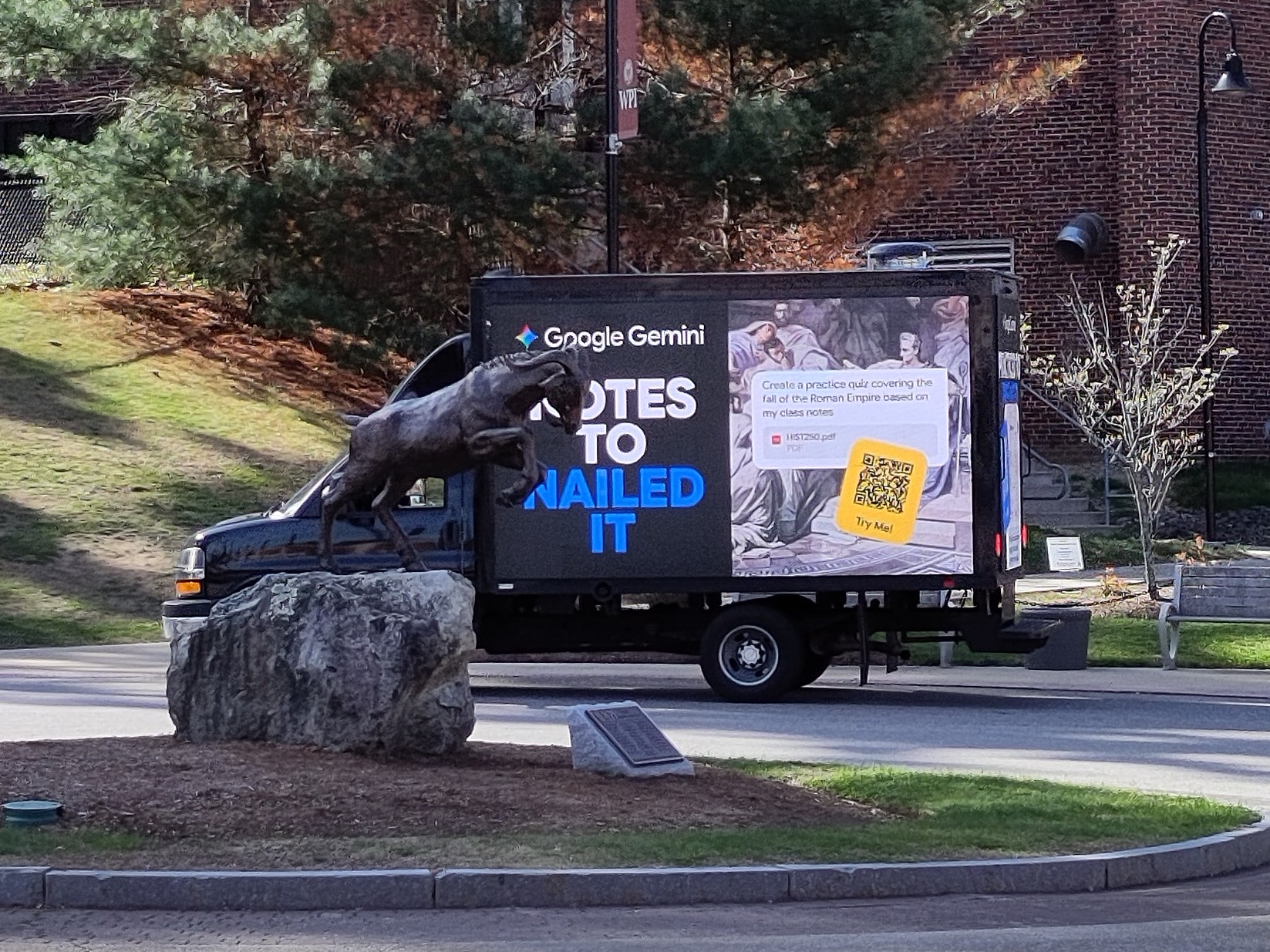 Black mobile billboard truck parked in cul-de-sac behind the WPI Sports and Recreation Center, in front of the pine tree between the back entrance to the Sports and Recreation Center and the service road leading to the Campus Center. The left billboard displays an advertisement with the headline "NOTES TO NAILED IT". In the center of the cul-de-sac is an island with a metal goat statue. The perspective makes it appear as if the goat is ramming the left billboard display with its horns.